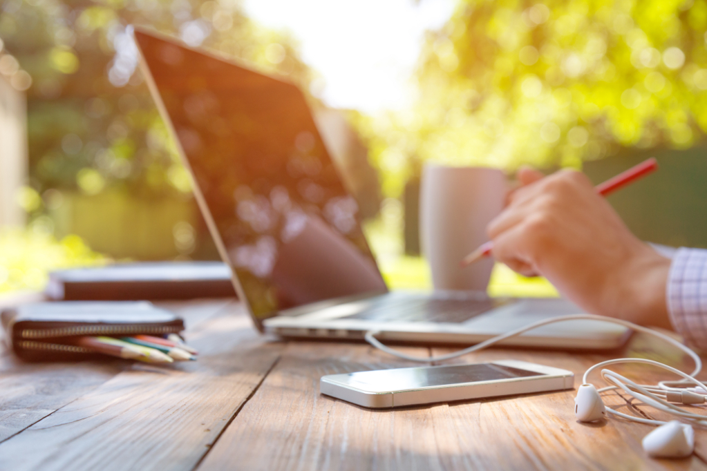 Casual dressed man sitting at wooden desk inside garden working on computer pointing with color pen electronic gadgets dropped around on table side view